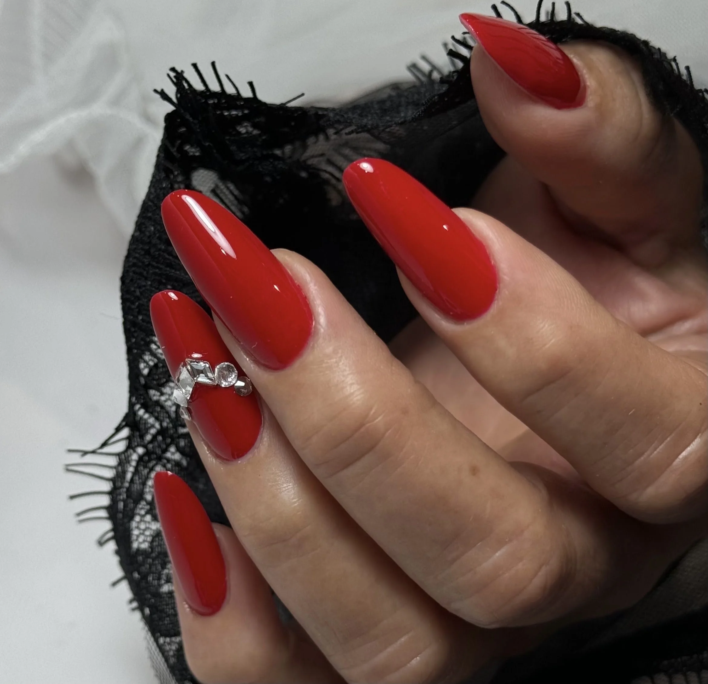 Close-up of a hand with red painted nails, wearing a ring with multiple large gemstones, holding black lace fabric.