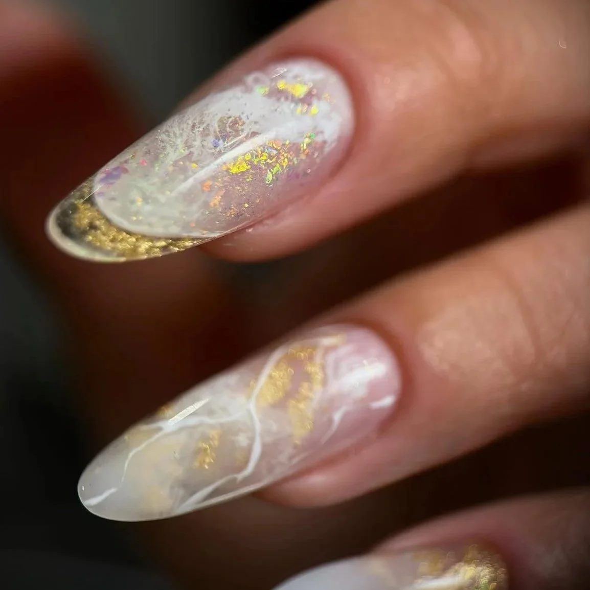 Close-up of a hand with decorated long, almond-shaped nails featuring a white, iridescent, and gold-flaked design.