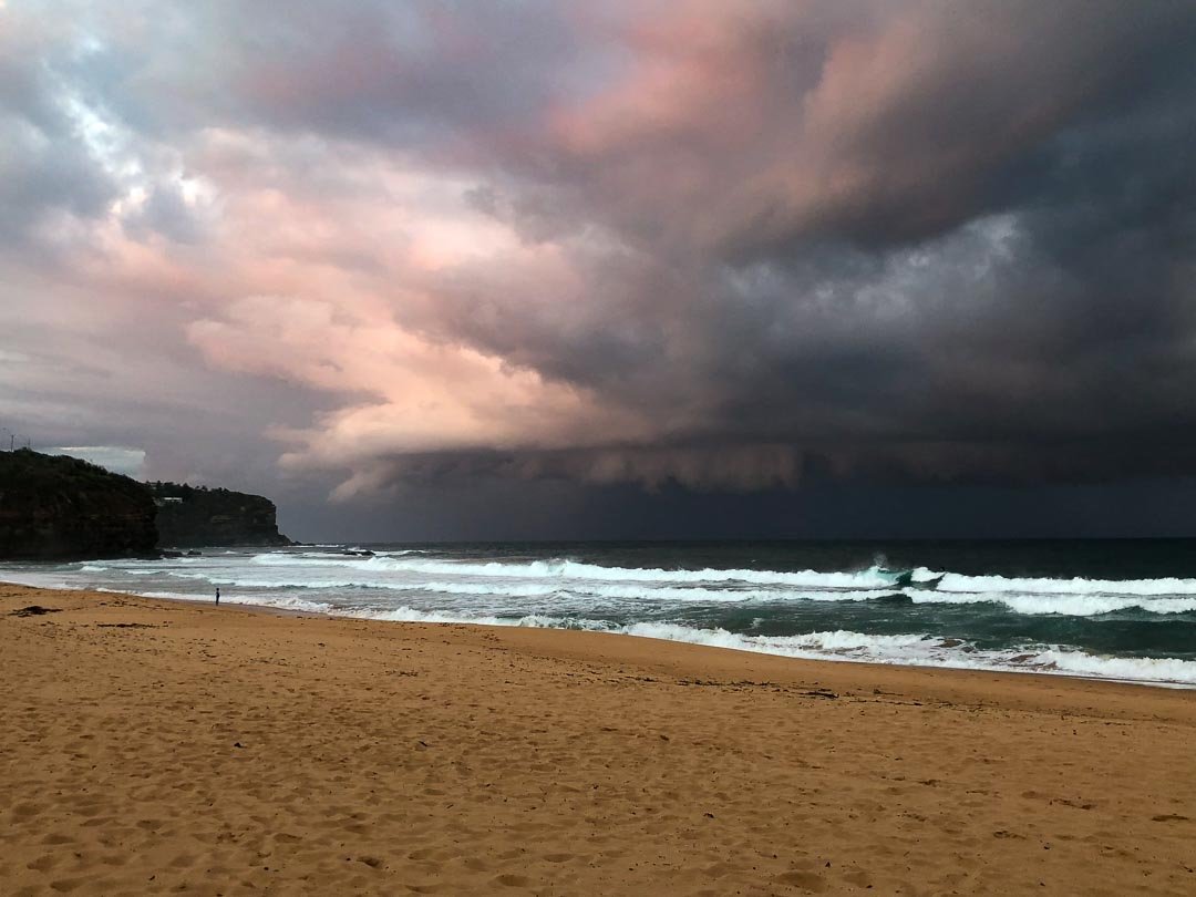 A beach with sandy shore and ocean waves under a dramatic sky with dark storm clouds and some pink and purple hues.
