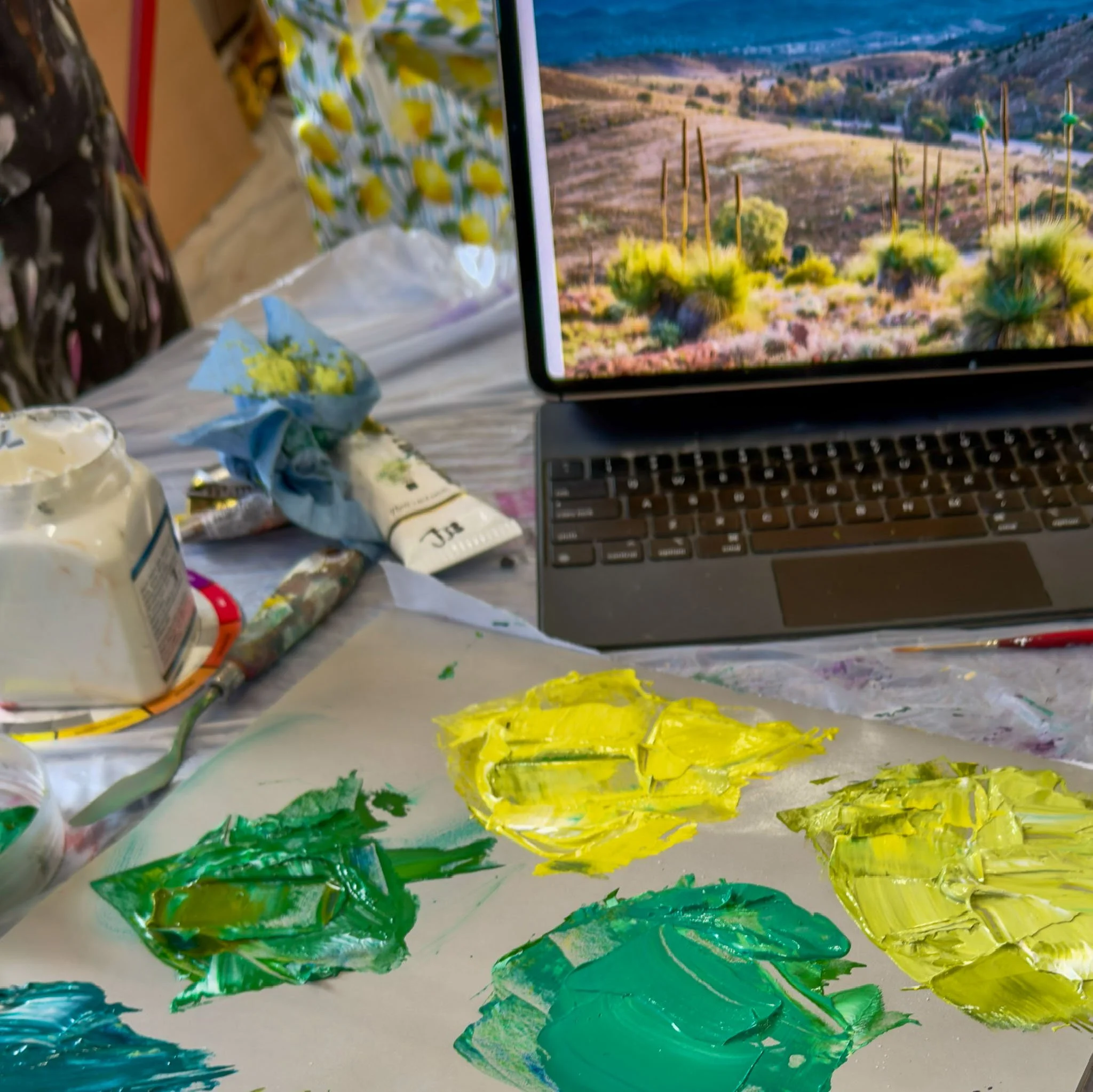 Close-up view of a paint palette with bright yellow, green, and blue acrylic paints, along with a partially visible paint tube and a laptop computer displaying a desert landscape with cacti in the background.