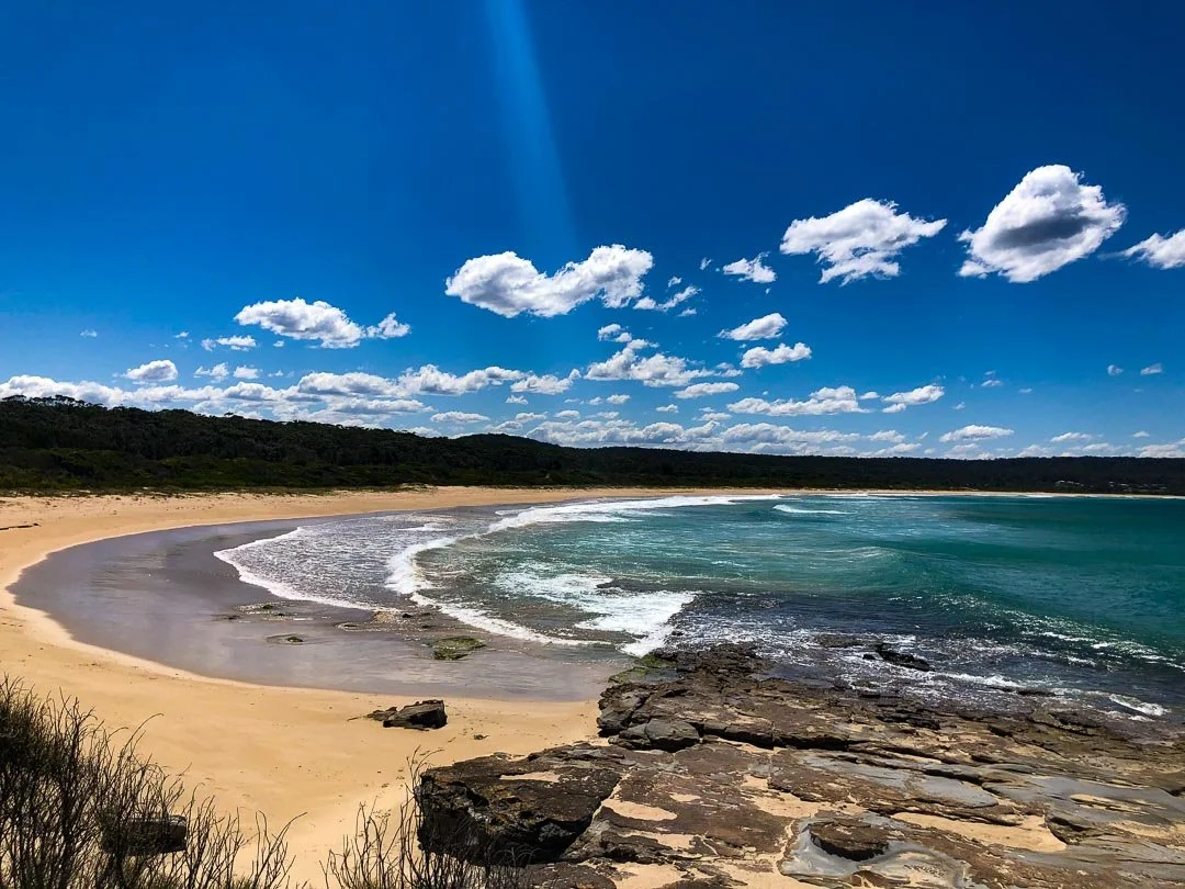 A scenic beach with golden sand, blue ocean waves, rocky outcrop in the foreground, and a green wooded hill in the distance under a partly cloudy sky.