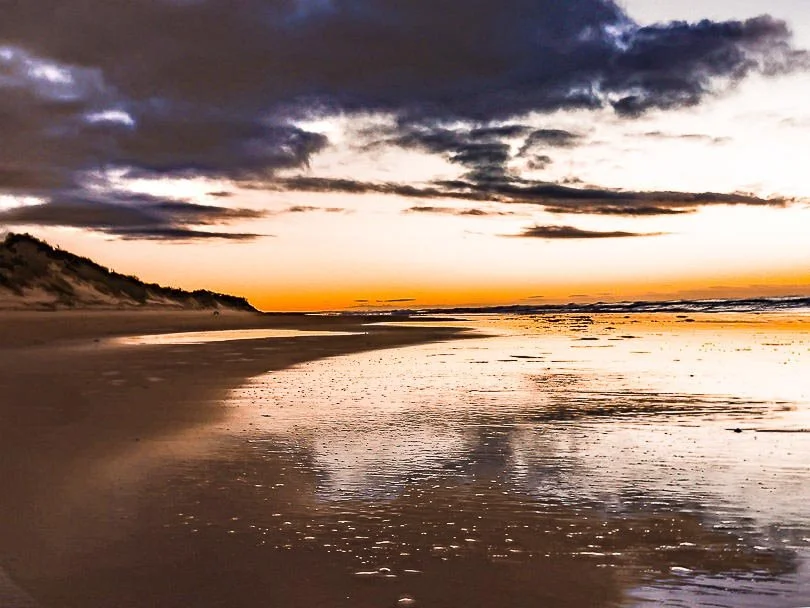 Sunset over a beach with wet sand and cloudy sky.