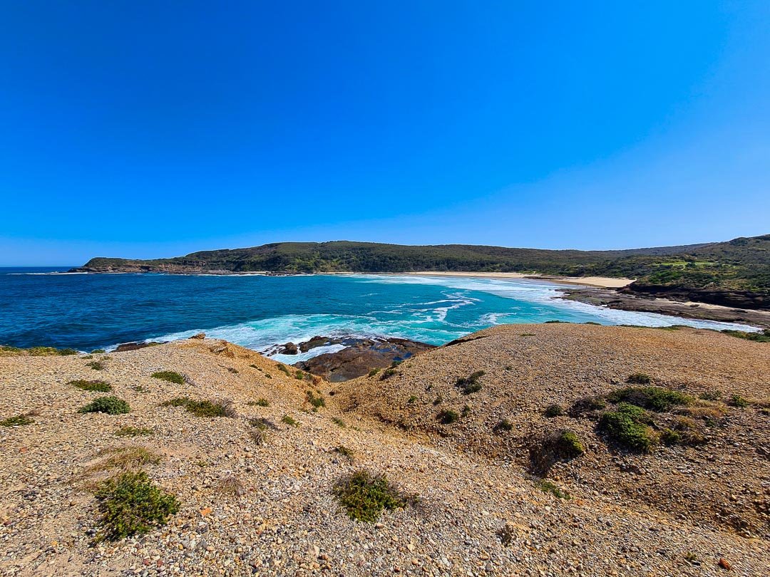 Coastal landscape with sandy cliffs, blue ocean waves, and a sandy beach under a clear blue sky.