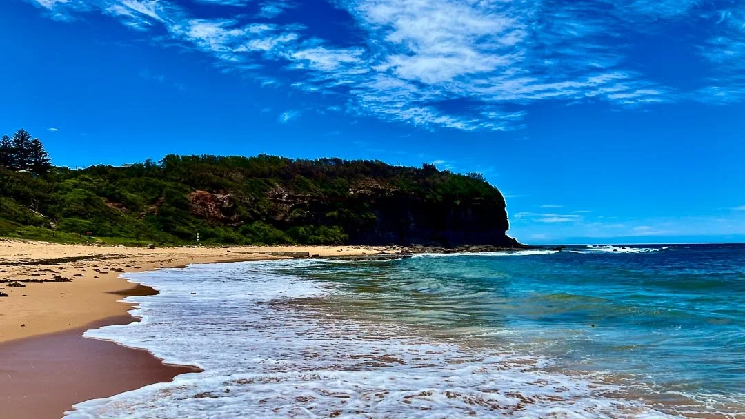 A scenic beach with golden sand, blue ocean waves, a green cliff in the background, and a partly cloudy sky.