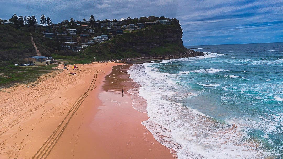 View of a sandy beach with waves, a cliff with houses on top, and a cloudy sky.