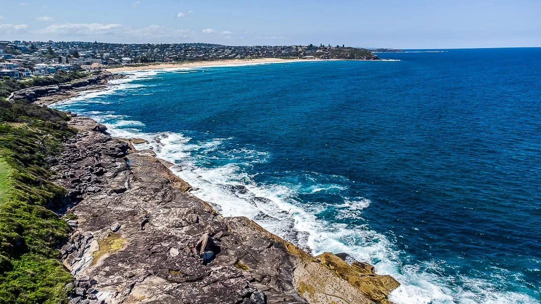 Overlooking a rugged coastline with rocks and greenery in the foreground, blue ocean waves crashing against the rocks, and a sandy beach in the distance under a partly cloudy sky.