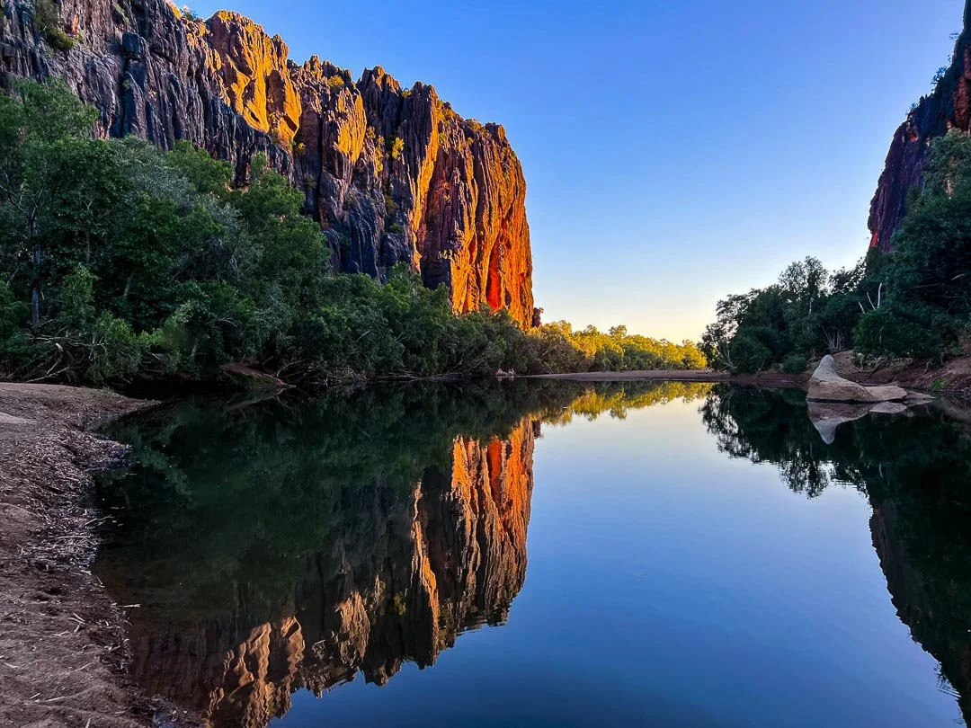 Sunlit cliffs reflecting in a calm river surrounded by green trees under a clear blue sky.
