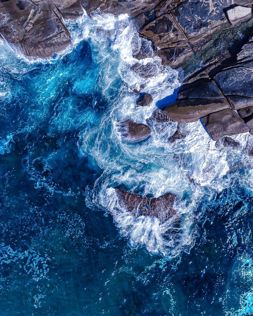 Aerial view of blue ocean waves crashing against large dark rocks along a shoreline.