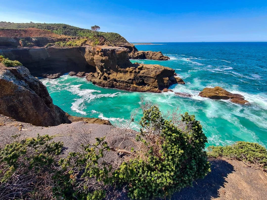 Cliffs overlooking the ocean with turquoise waves crashing against rocks on a sunny day.