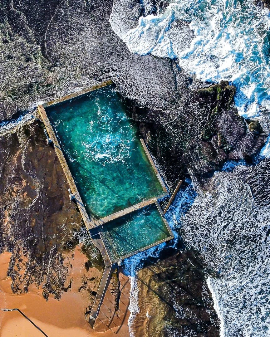 Aerial view of a large ocean pool built on rocky terrain, with waves crashing nearby and a sandy beach at the bottom left corner.