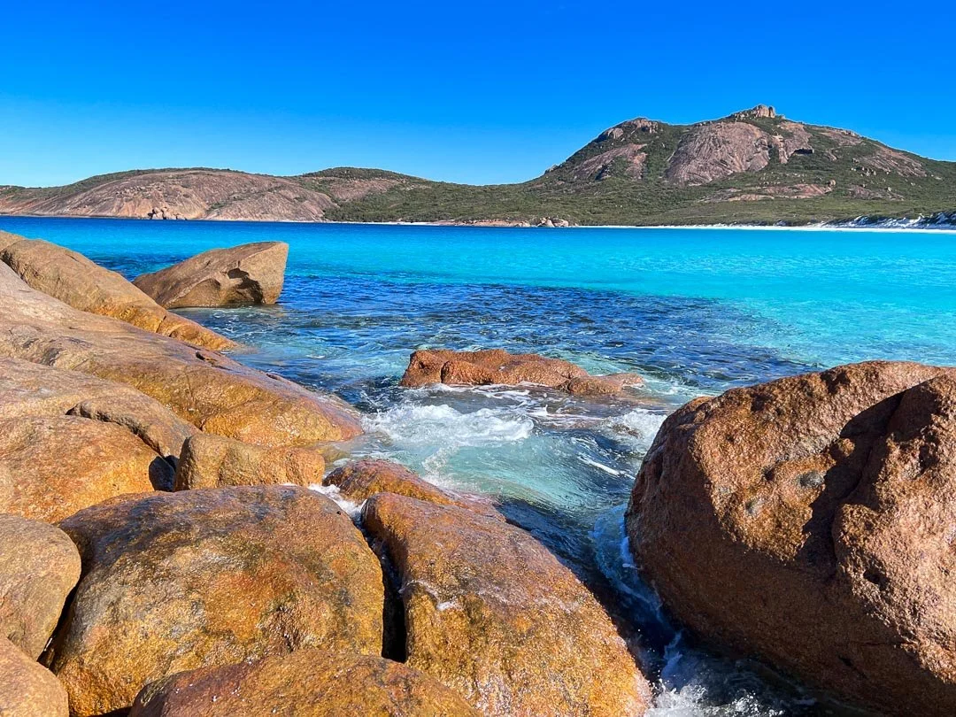 Clear blue ocean with large rocks in the foreground and a mountainous landscape in the background under a bright blue sky.