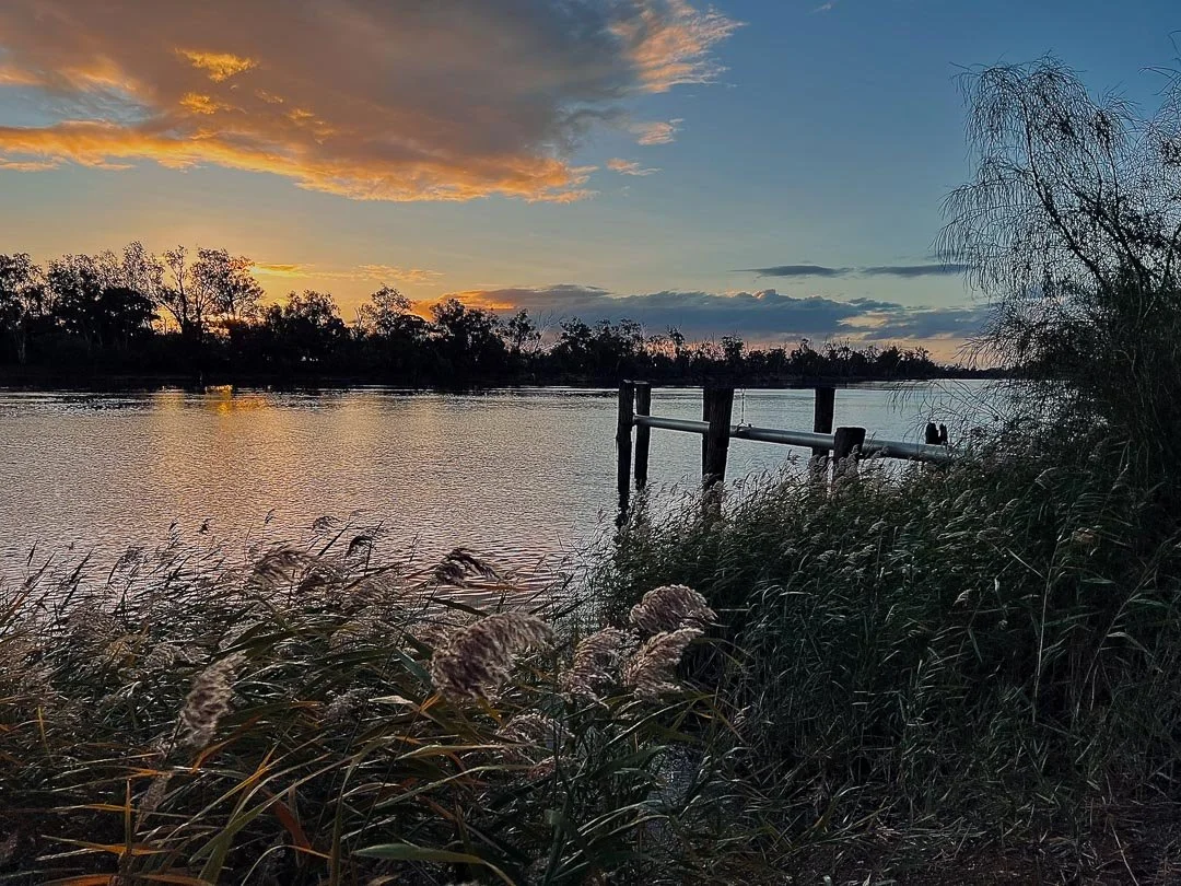 A serene river scene at sunset with a partly cloudy sky, silhouettes of trees along the horizon, reeds in the foreground, and a small dock extending into the water.