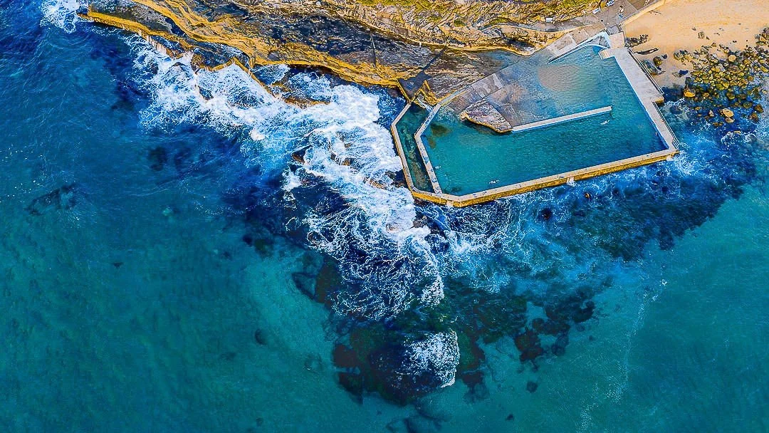 A small ocean pool surrounded by rocky cliffs, with waves crashing over the rocks and flowing into the pool.