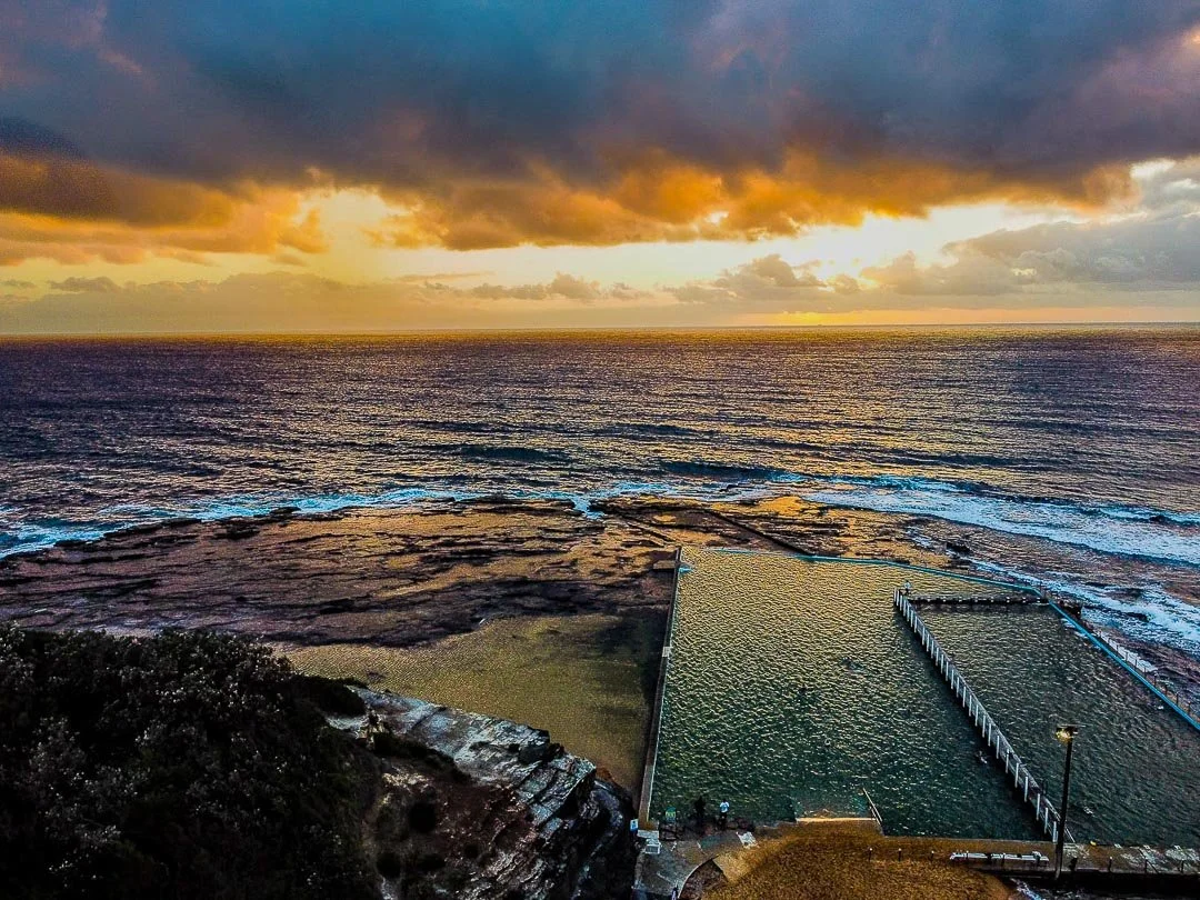 A sunset over the ocean with a tidal pool and rocky shore in the foreground.