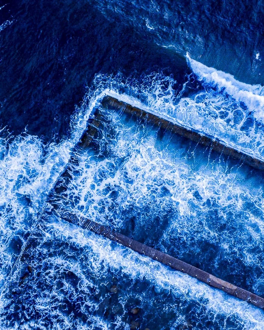 Aerial view of turbulent blue ocean waves crashing over a concrete breakwater or jetty.