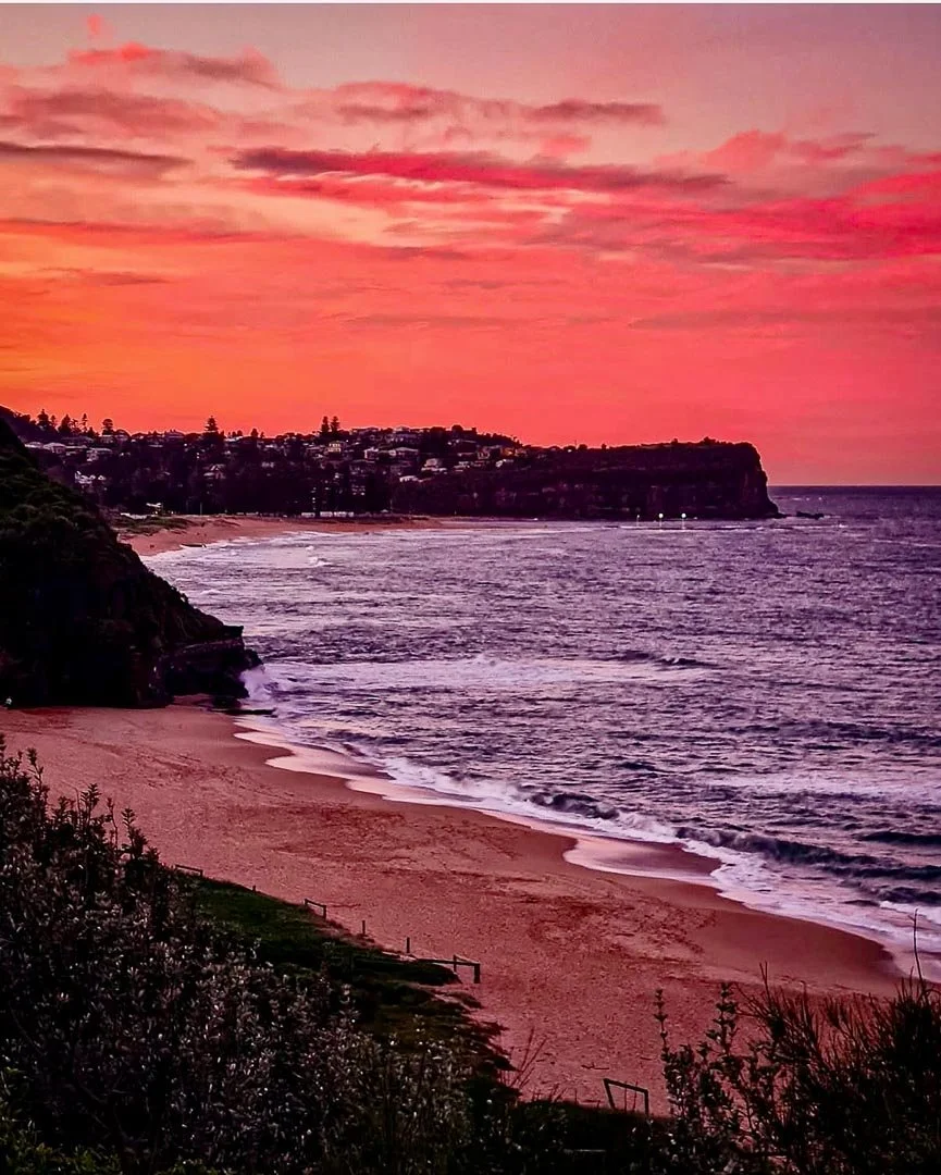 Sunset over a sandy beach with waves, rocky cliff on the left, and houses along the shoreline in the distance.
