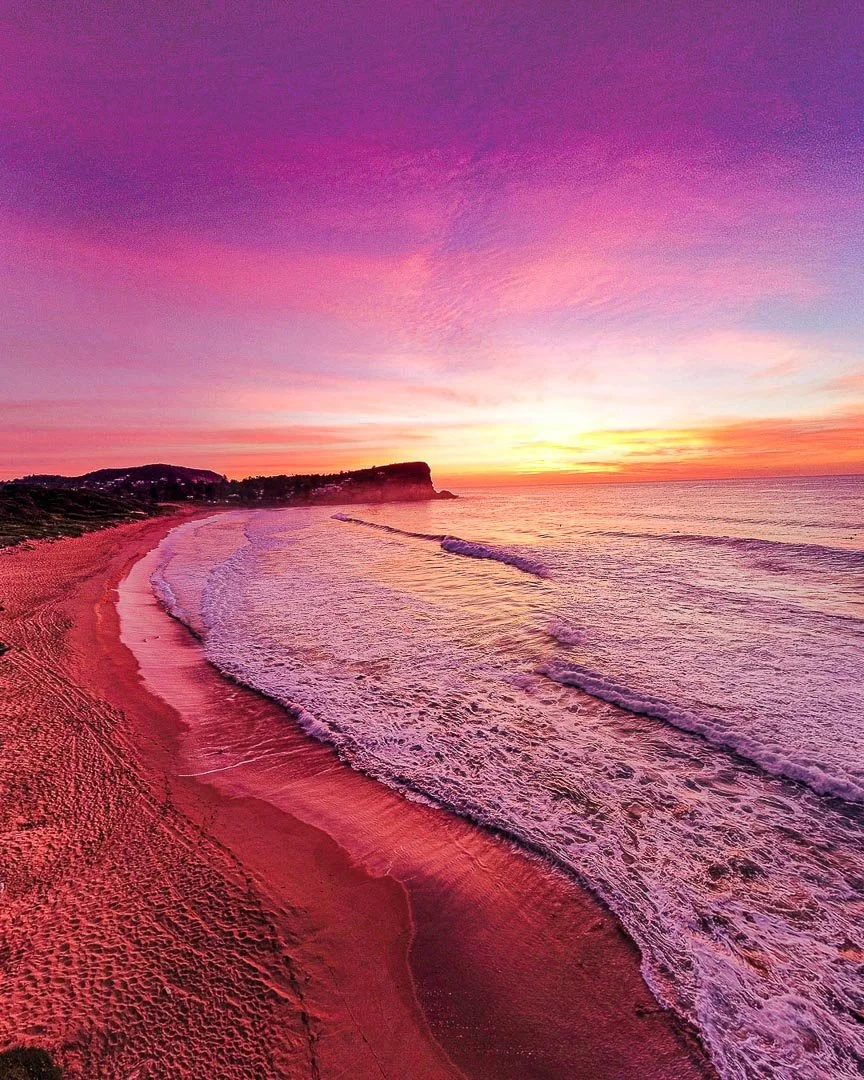 A colorful sunset over a beach with pink and purple sky, gentle waves, and shoreline.