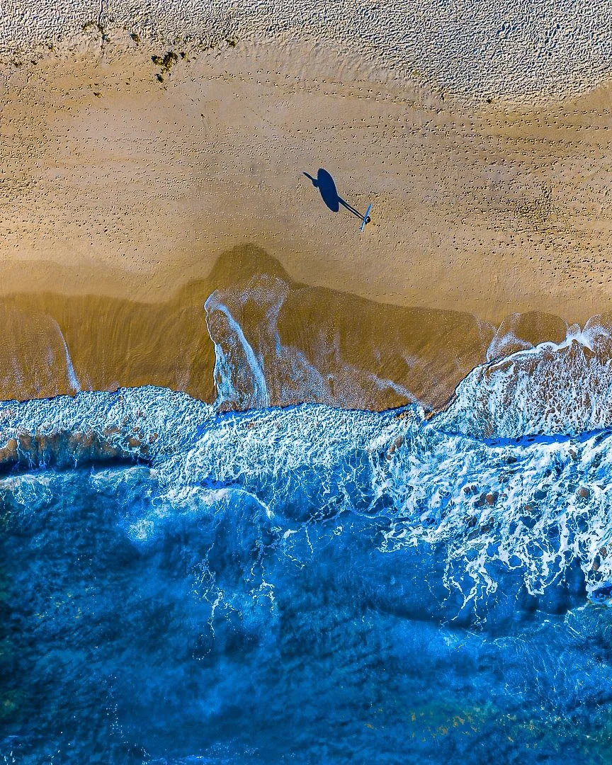 An aerial view of a beach with ocean waves crashing onto the shore, seen from above. A person is walking on the sandy beach creating footprints.