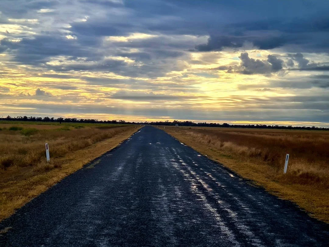 A long, straight asphalt road extending into the horizon through a rural landscape, with golden field grass on either side and a partly cloudy sky overhead with sunlight breaking through.