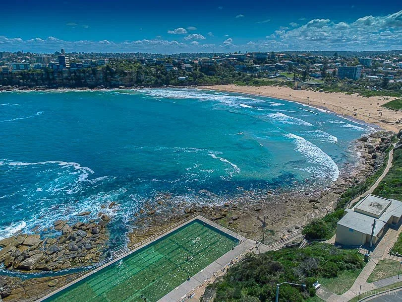 A scenic aerial view of a beach with turquoise water, a rocky shoreline, and a sandy beach area with people. In the foreground, there is a rectangular swimming pool, and in the background, a city skyline with buildings and greenery under a partly clo