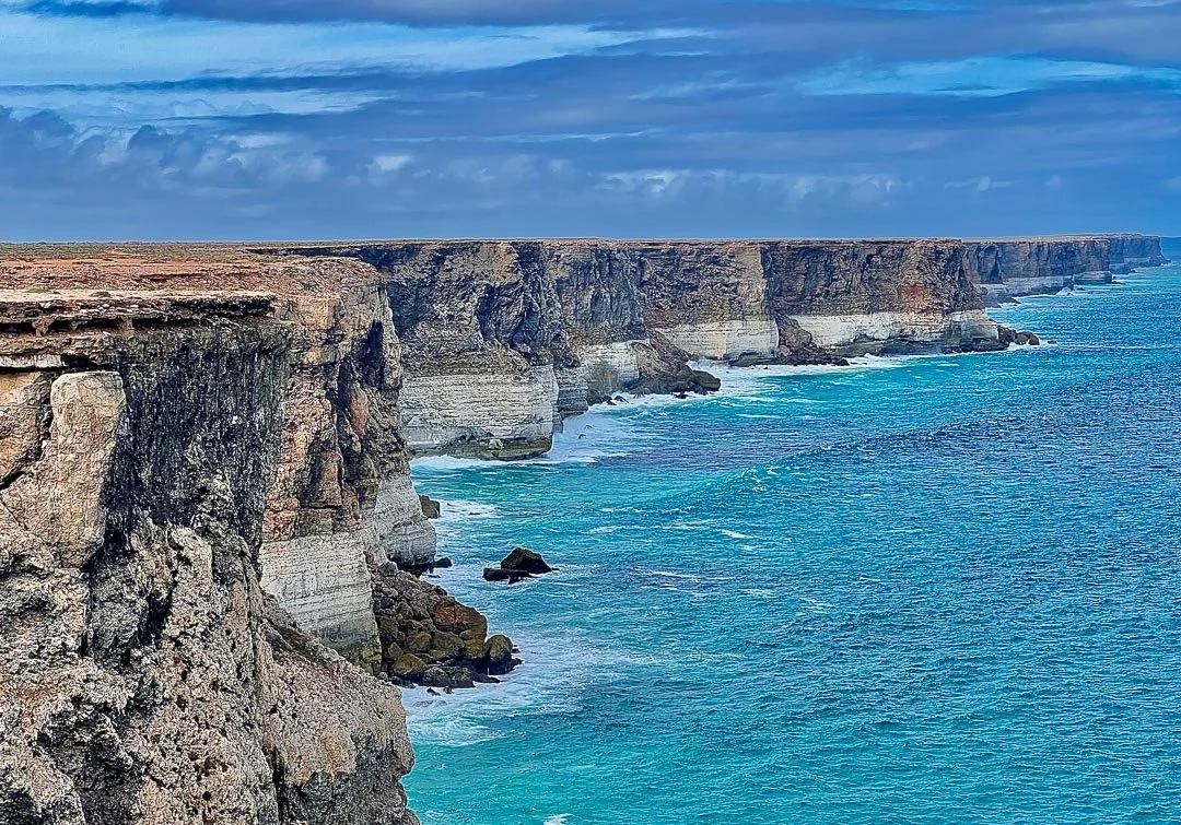 Cliffs along a rugged coastline with white waves crashing at the base, under a partly cloudy sky.