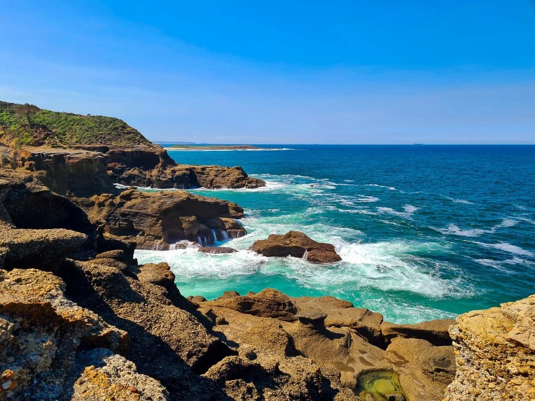 A scenic coastal landscape with rocky cliffs, blue ocean waves, and a clear blue sky.