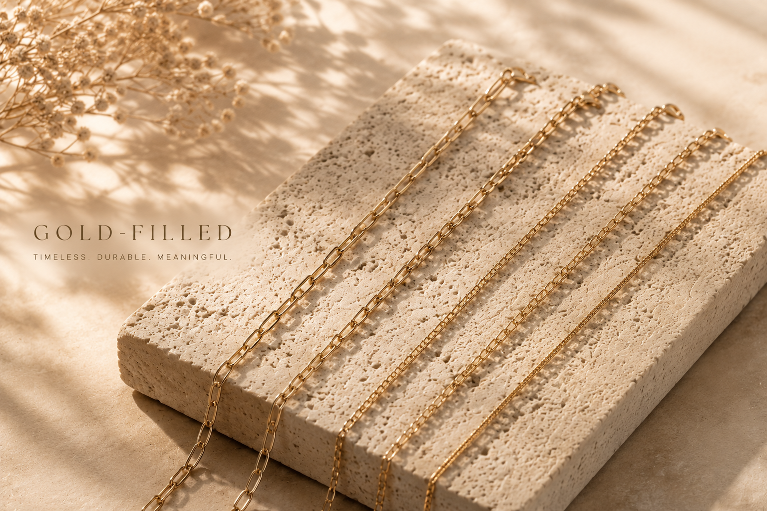 Six gold-filled necklaces resting on a textured white stone slab, with a sprig of dried white flowers to the left.