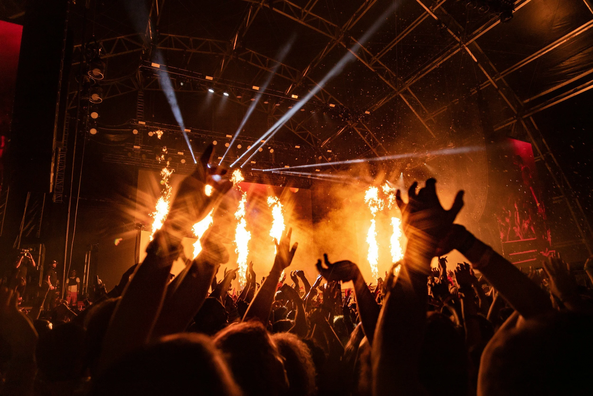 Crowd at a concert or festival with hands raised, fire and pyrotechnics on stage, bright beams of lights, and a large indoor stage structure.