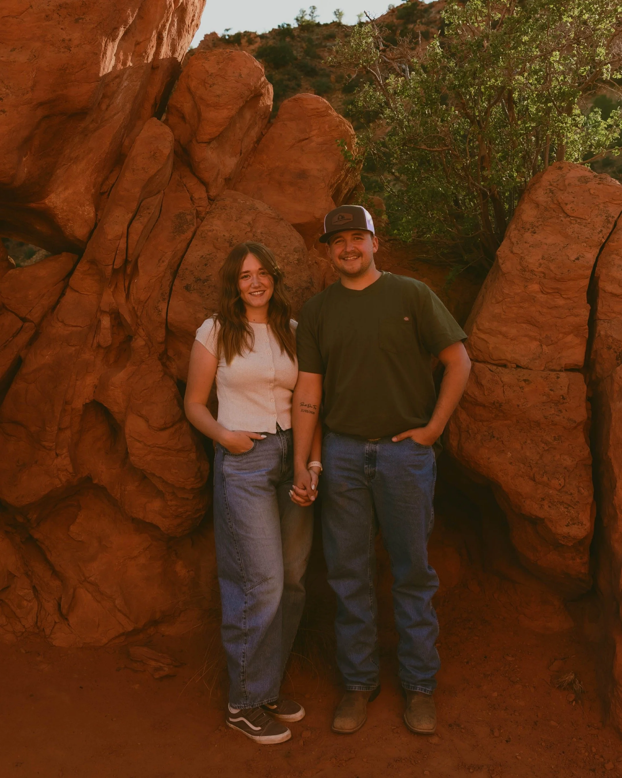 A smiling couple stands close together, holding hands, in front of large red rock formations and green trees in a desert landscape on a sunny day.