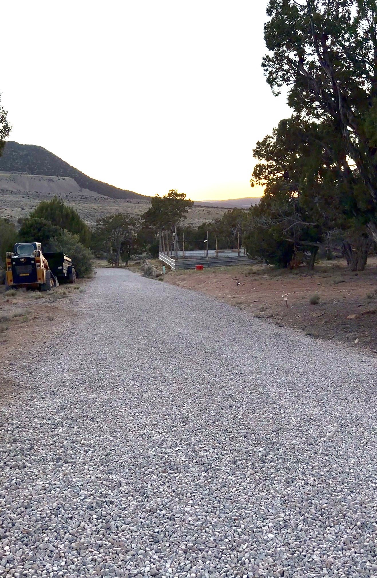 A gravel road running through a rural area with trees and a mountain in the background, and a construction vehicle parked to the left.