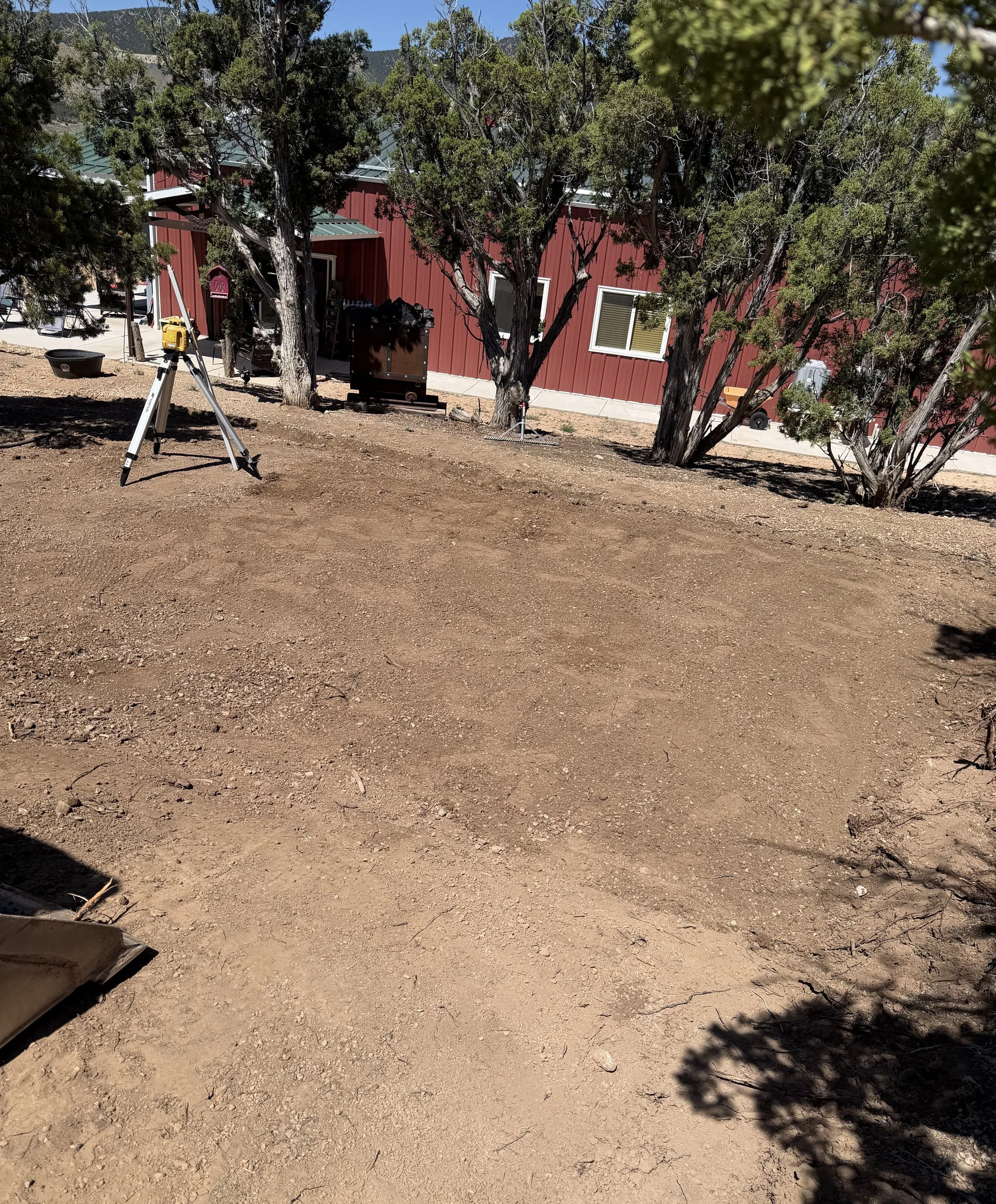 A dirt yard with shadows of trees in the foreground, a red building with white window frames and a metal roof, and various outdoor items including a wheelbarrow, a trash bin, and a yellow device on a tripod.