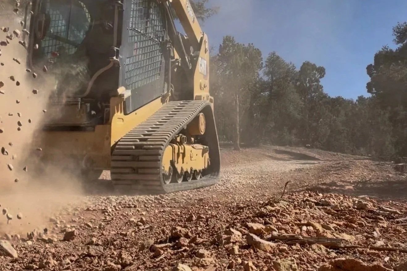 A yellow bulldozer moves earth and rocks on a dirt trail surrounded by trees under a partly cloudy sky.