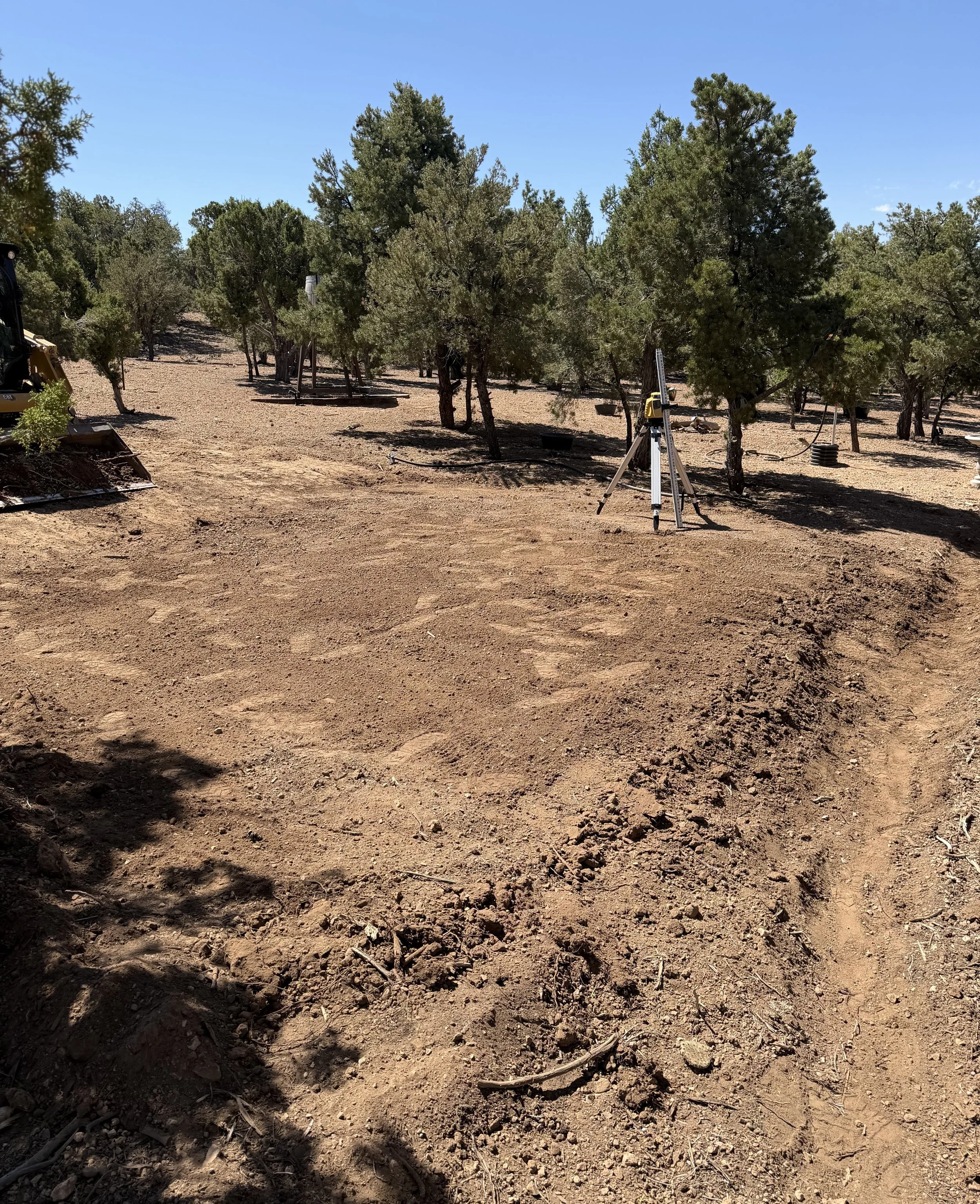 A cleared dirt area with scattered small rocks and organic debris under a clear blue sky, with several trees and some pieces of equipment or structures in the background.