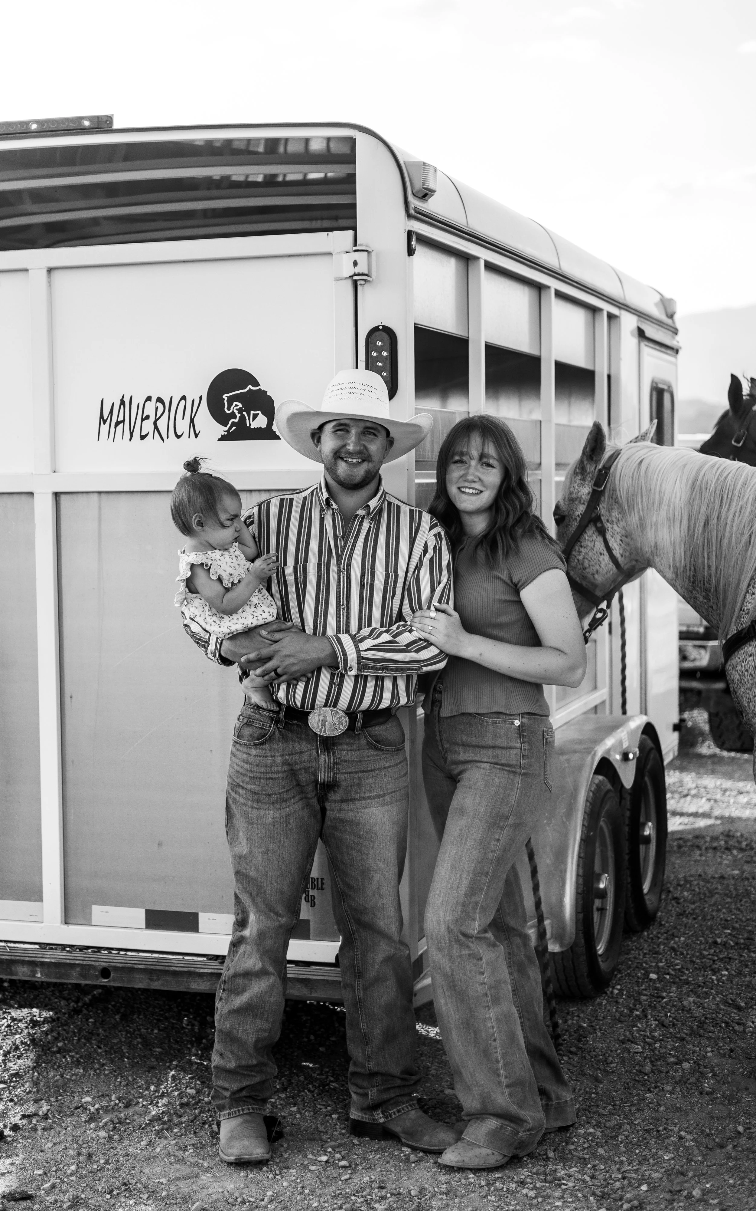 A man wearing a cowboy hat and a striped shirt, a woman in a short-sleeved shirt, and a young girl in a floral dress stand together in front of a horse trailer titled "MAVERICK." The man is holding the girl, and the woman has her arm around the man's waist. There is a horse visible to the right side of the image.