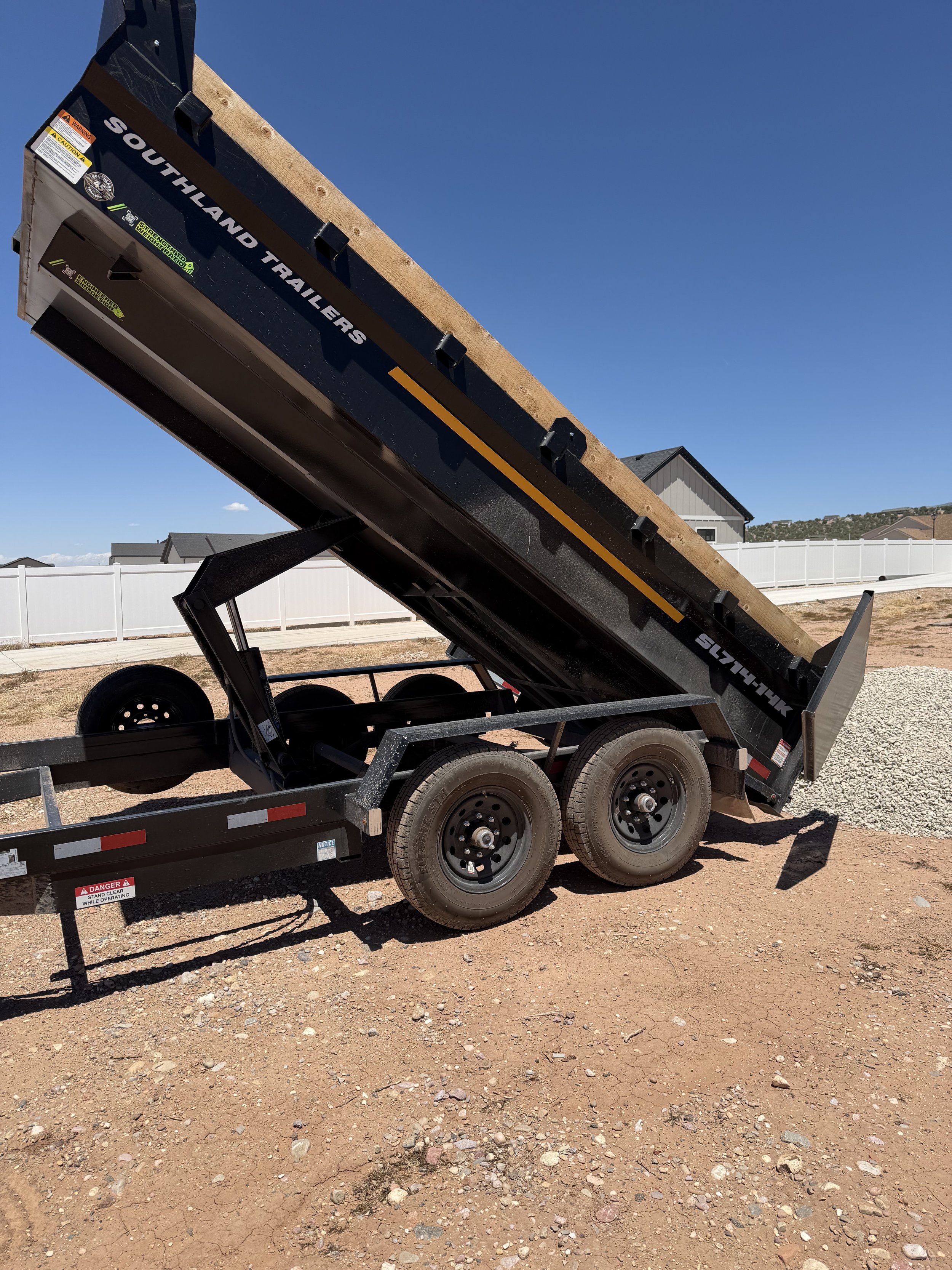 A black trailer with tires, lifting a wooden board marked 'SOUTHLAND TRAILERS' against a clear blue sky, with a white fence, a house, and mountains in the background.