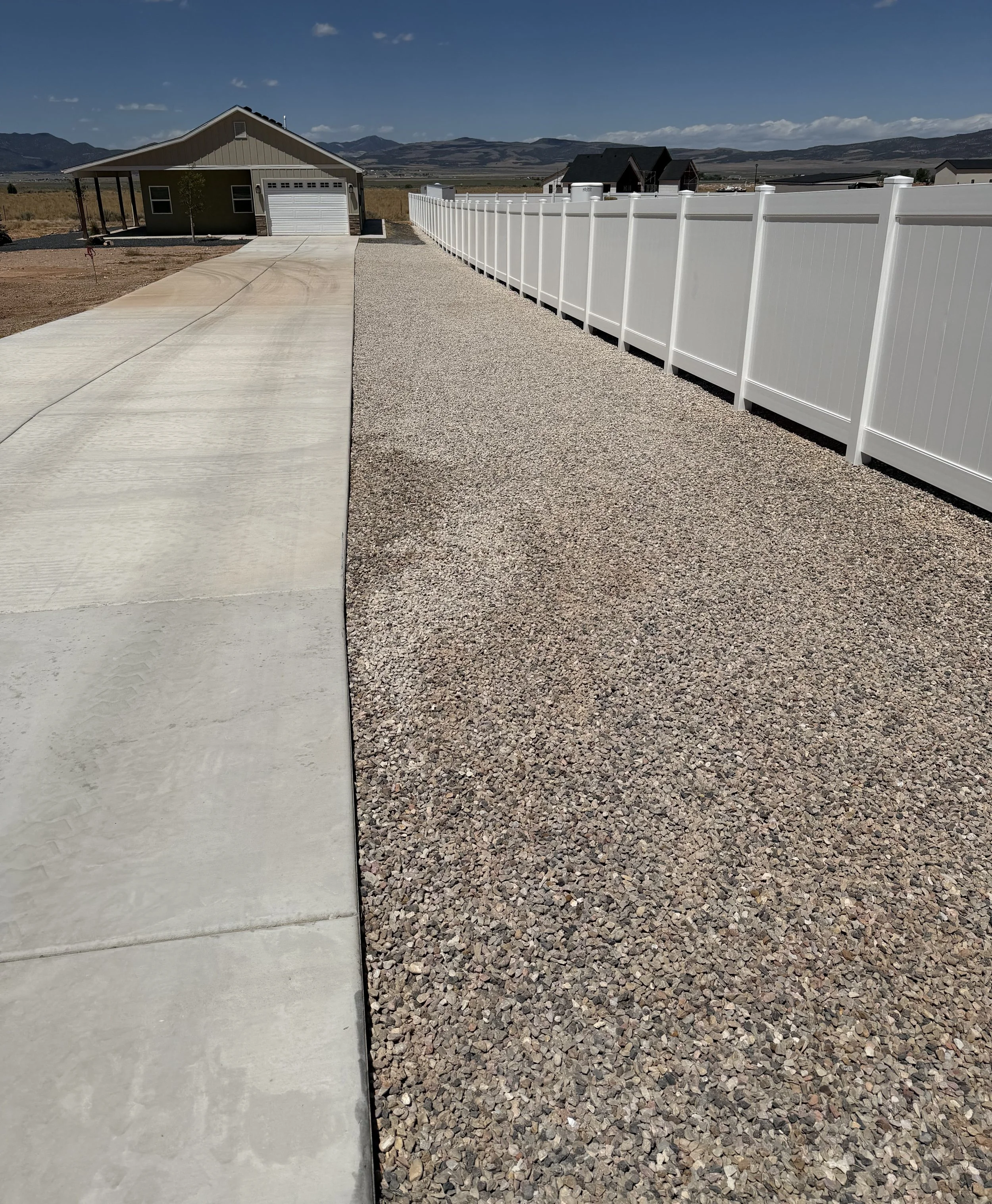A driveway leading to a garage with a house on the left and a white vinyl fence on the right, set in a rural area with mountains in the background under a blue sky with a few clouds.