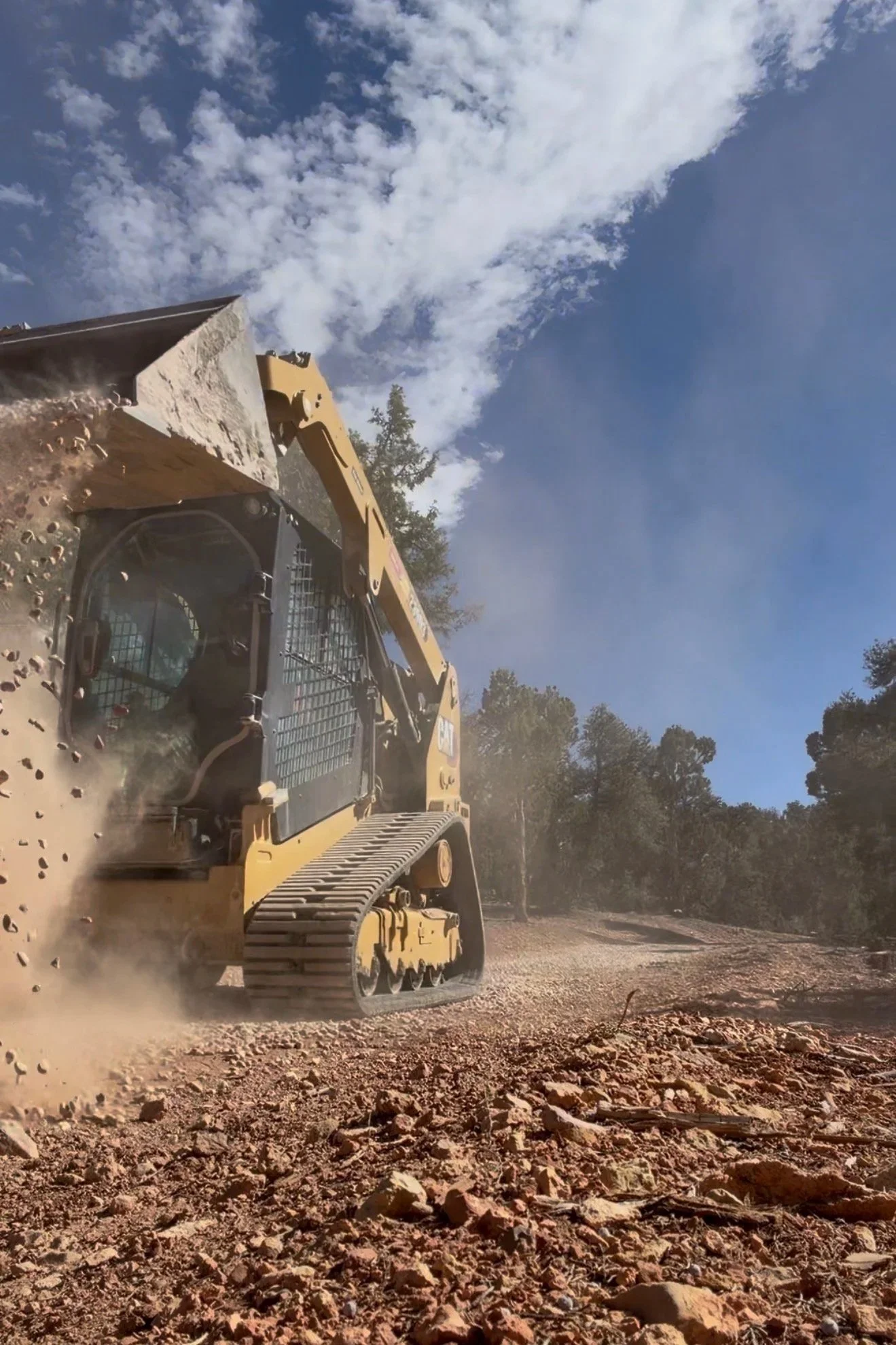 A yellow bulldozer working on a dirt road under a partly cloudy sky, with trees in the background.
