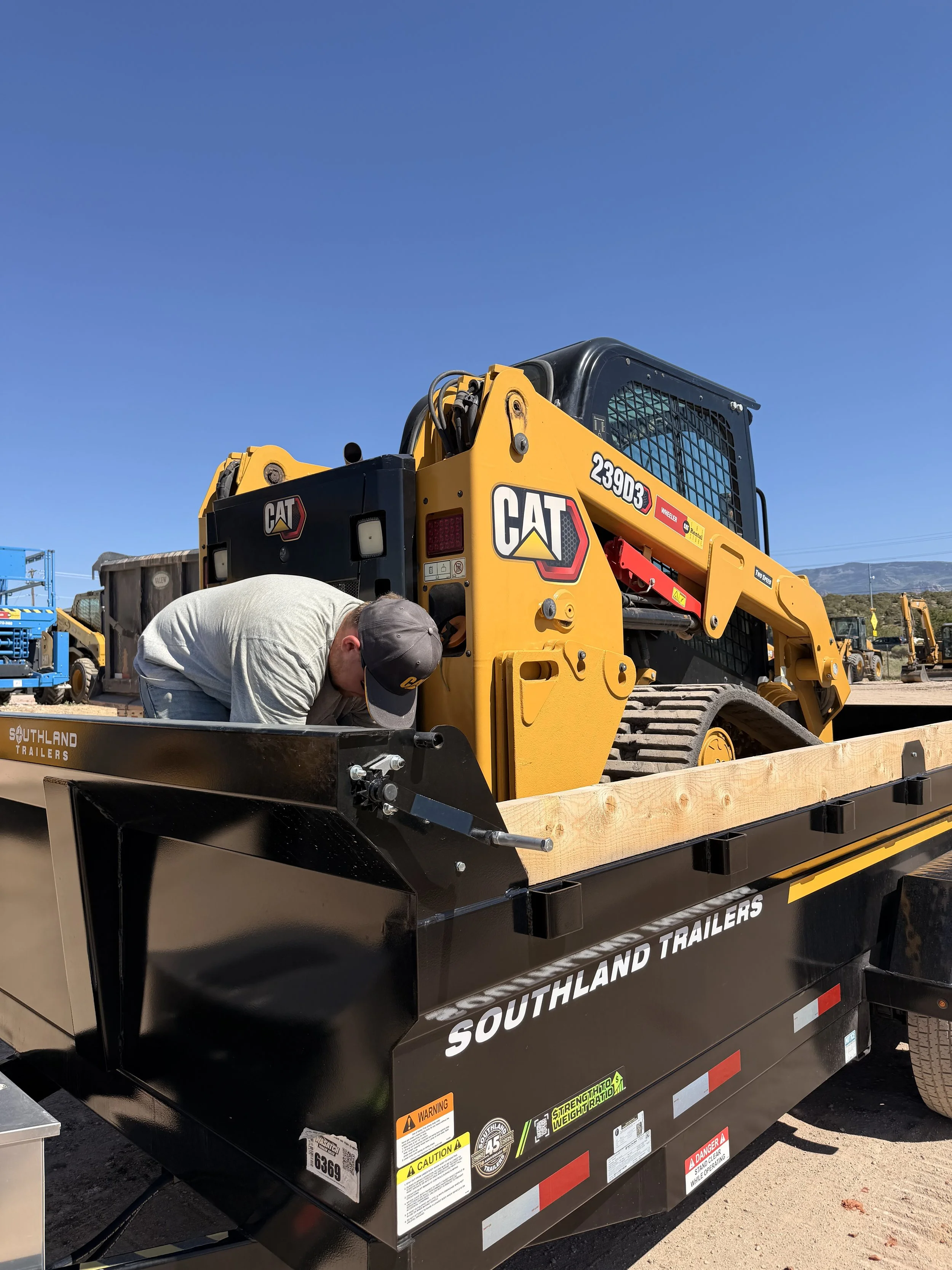 A construction worker is leaning over a black trailer with the Southland Trailers logo, next to a yellow Caterpillar compact track loader. Clear blue sky in the background.