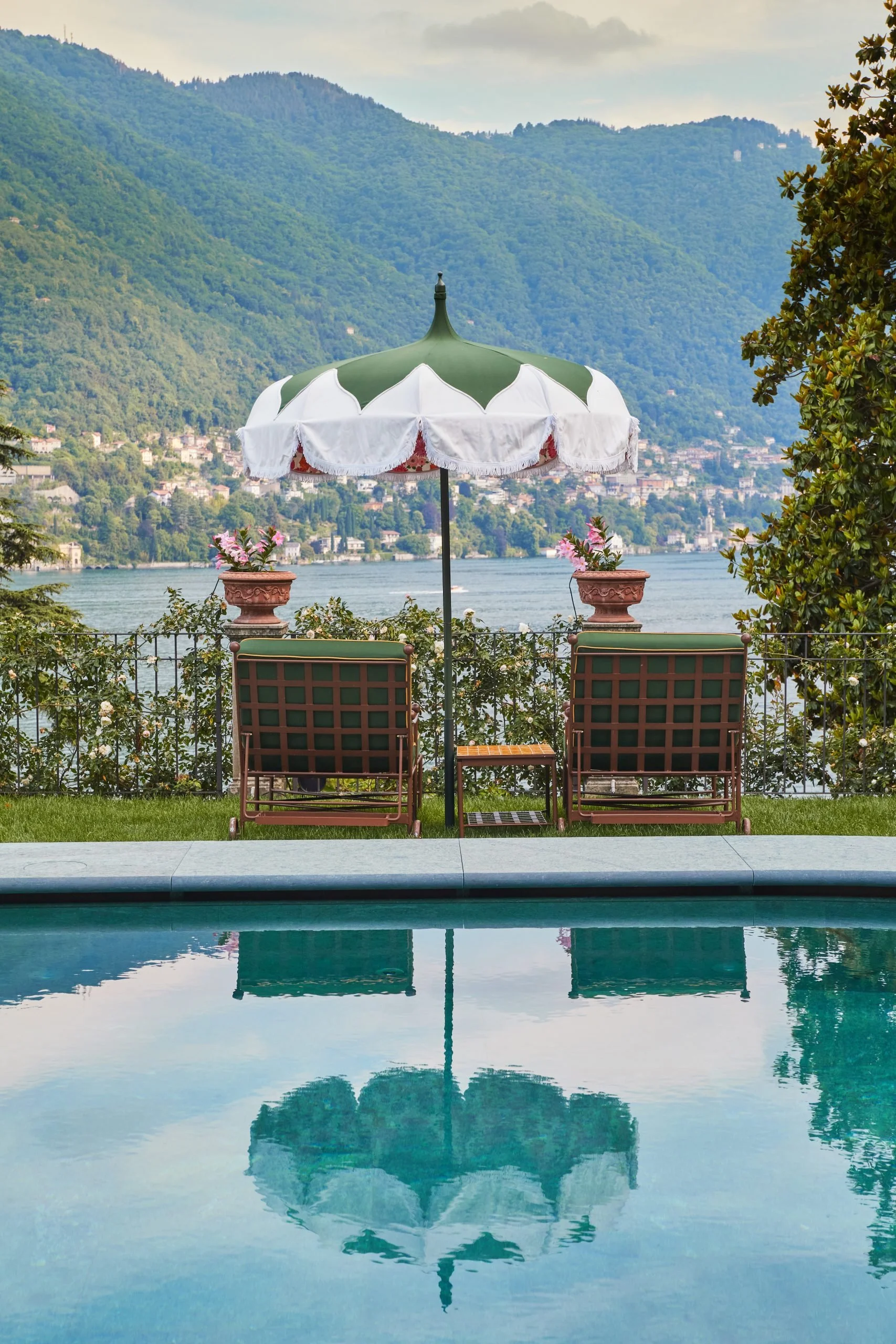 A view of a poolside scene with two brown patio chairs and a green and white umbrella. In the background, there are two flower pots with pink flowers, a river, lush green mountains, and a partly cloudy sky.