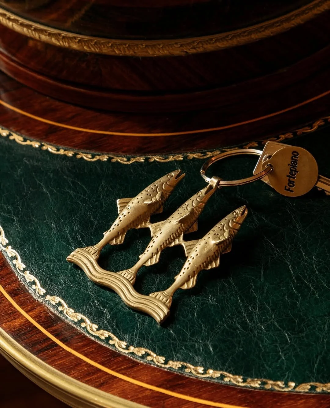 Gold-colored keychain with three fish shaped charms on a green leather surface with gold decorative trim, and a wooden object in the background.