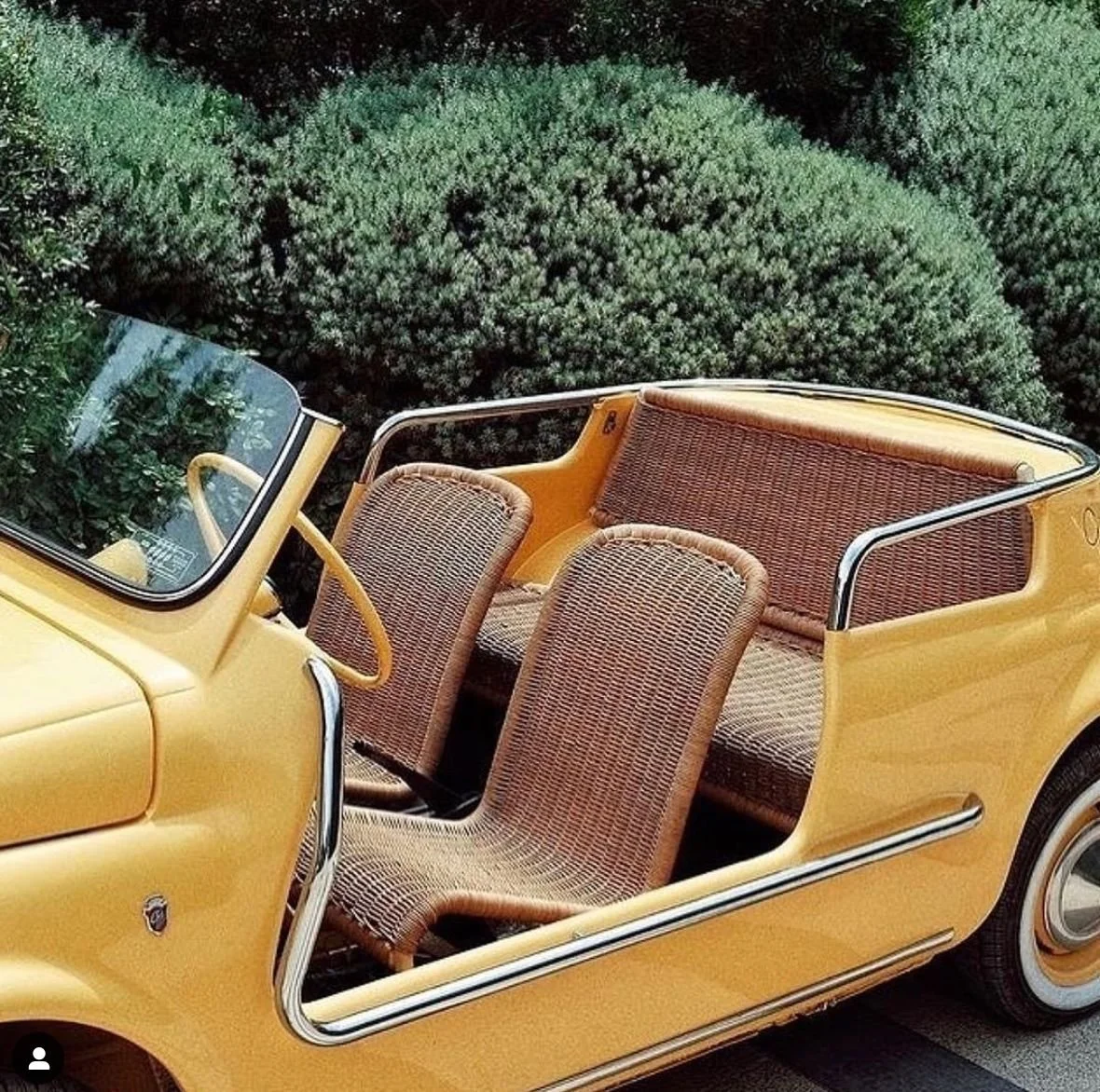 A vintage yellow autovolkswagen with wicker seats and a convertible top parked in front of green bushes.