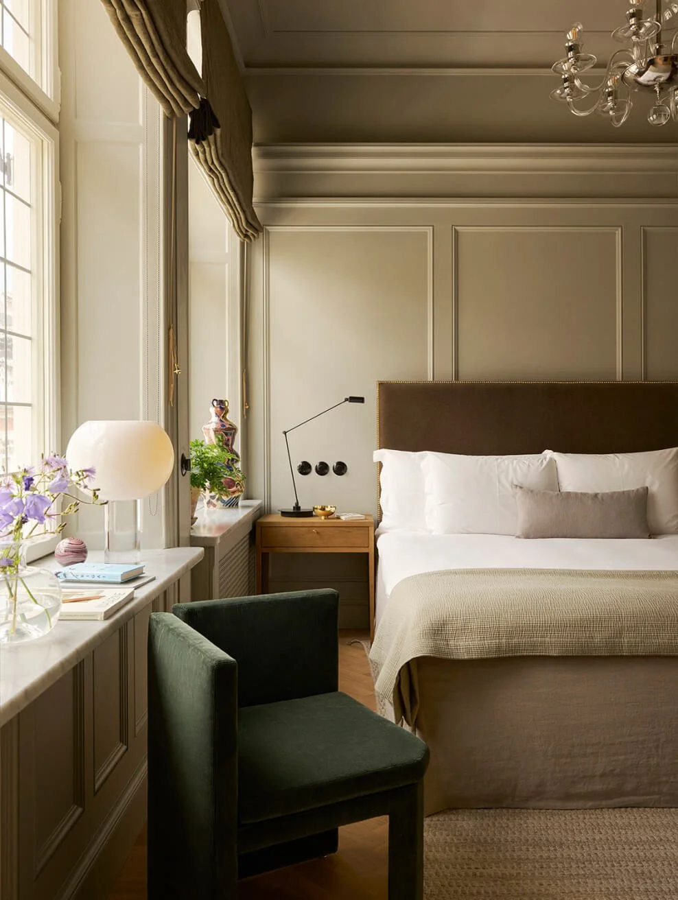 Elegant bedroom with large window, beige walls, green velvet chair, wooden nightstand with lamp, decorative vase, and bed with white linens and and beige bedspread.