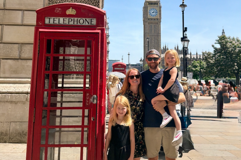 A family of four with two young girls standing next to a red London telephone booth. The father is holding one girl, and the mother and the other girl are standing beside him. In the background, Big Ben and the Houses of Parliament are visible, with other tourists and street vendors around.