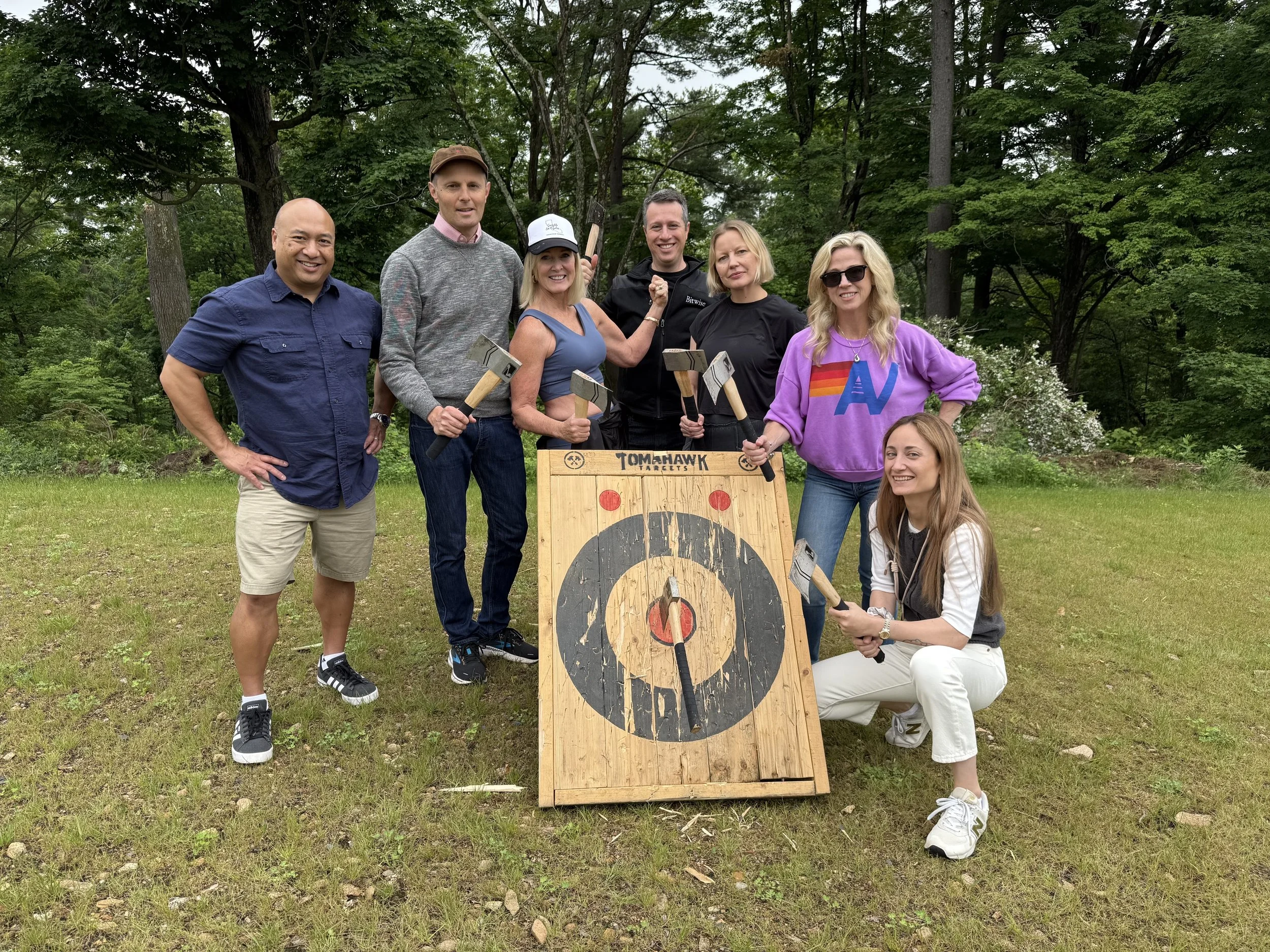 Group of seven diverse adults outdoors in a wooded area, smiling and holding axe handles around a wooden Target targets game with a large target board, some kneeling and others standing, with trees and greenery in the background.