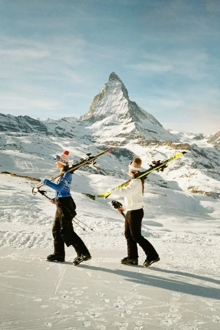 Two people in ski gear walking on snow with skis on their shoulders, Pike's Peak Mountain in the background.