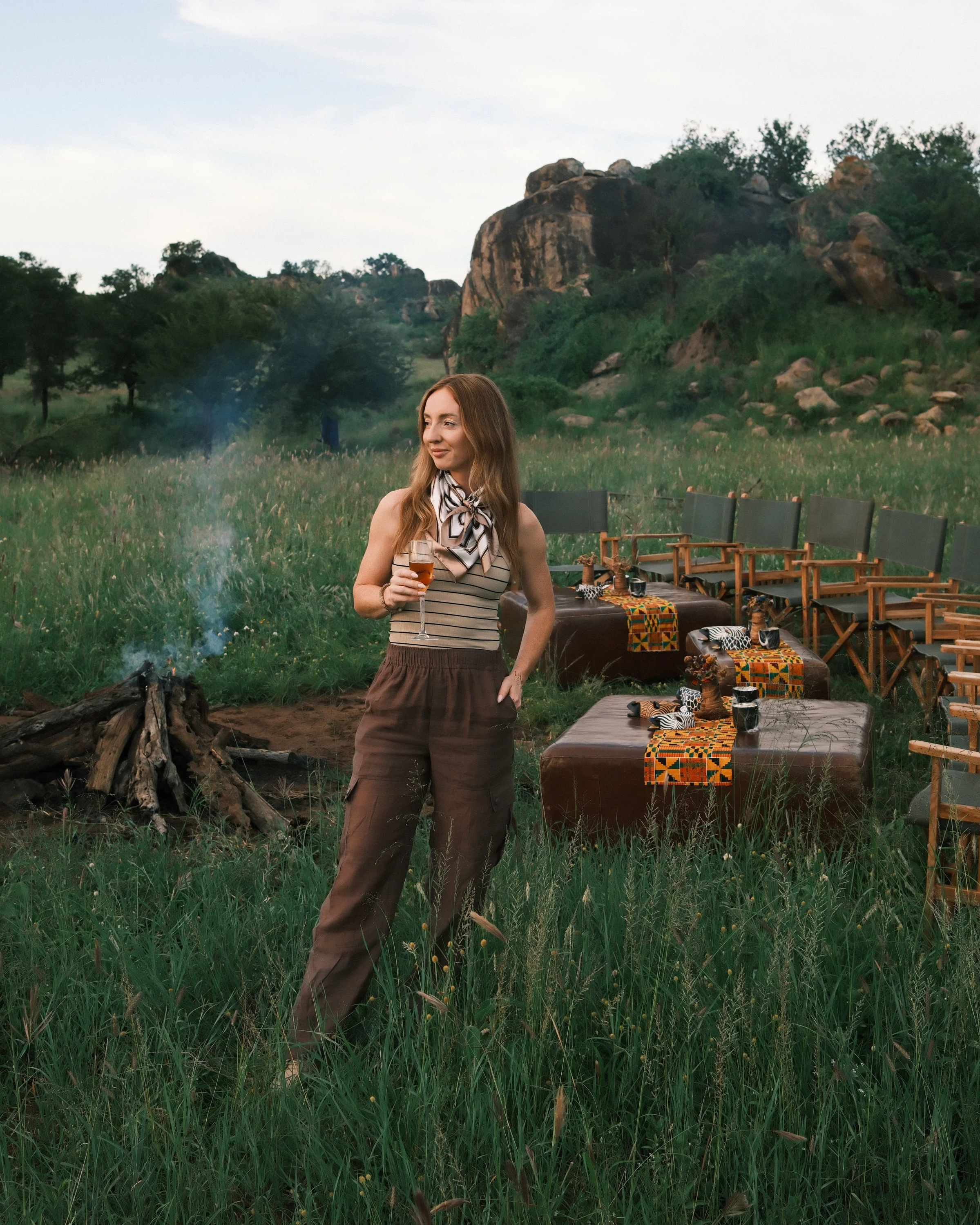 A woman with long red hair standing in a grassy field holding a glass of wine, with outdoor chairs and a table with a colorful table runner behind her. The background features green hills, trees, and large rocks, suggesting a natural outdoor setting during the daytime.