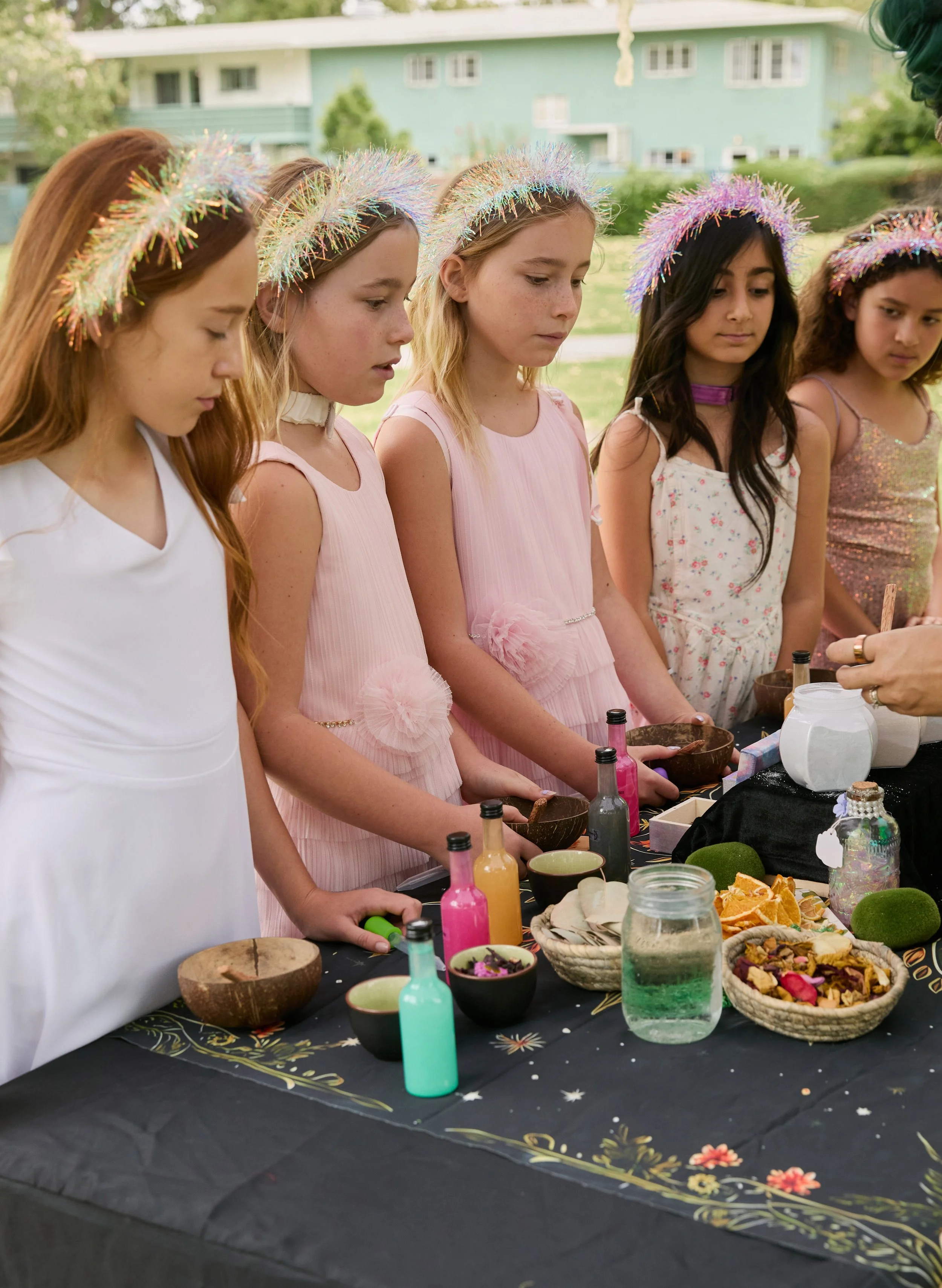 A group of young girls in fairy dresses and shiny headbands gathered around a table outdoors, observing a potion-making activity