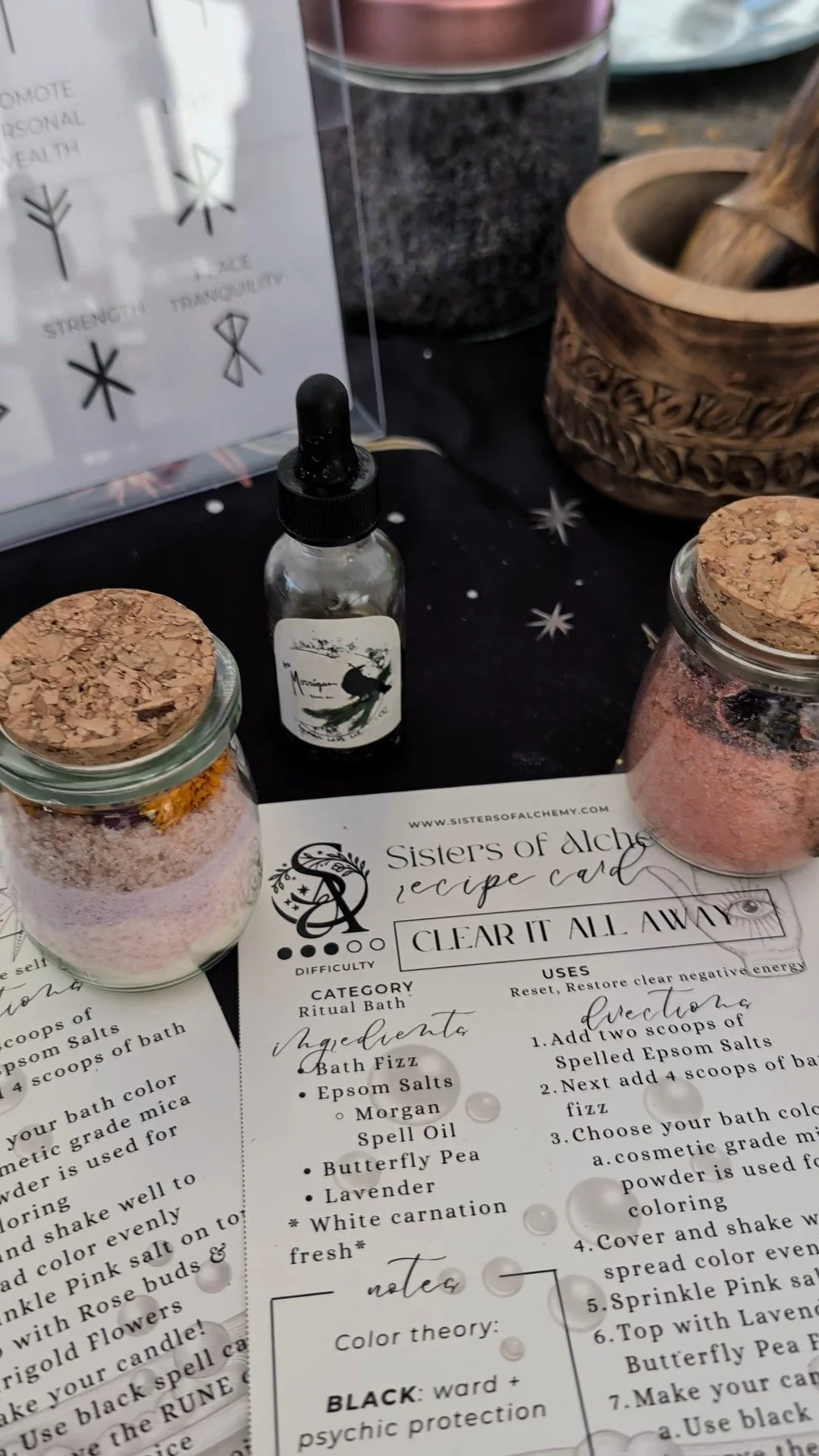 Sisters of Alchemy spiritual wellness ritual kit featuring handmade pink bath salts in corked glass jars, an essential oil dropper bottle, and a Clear It All Away ritual recipe card