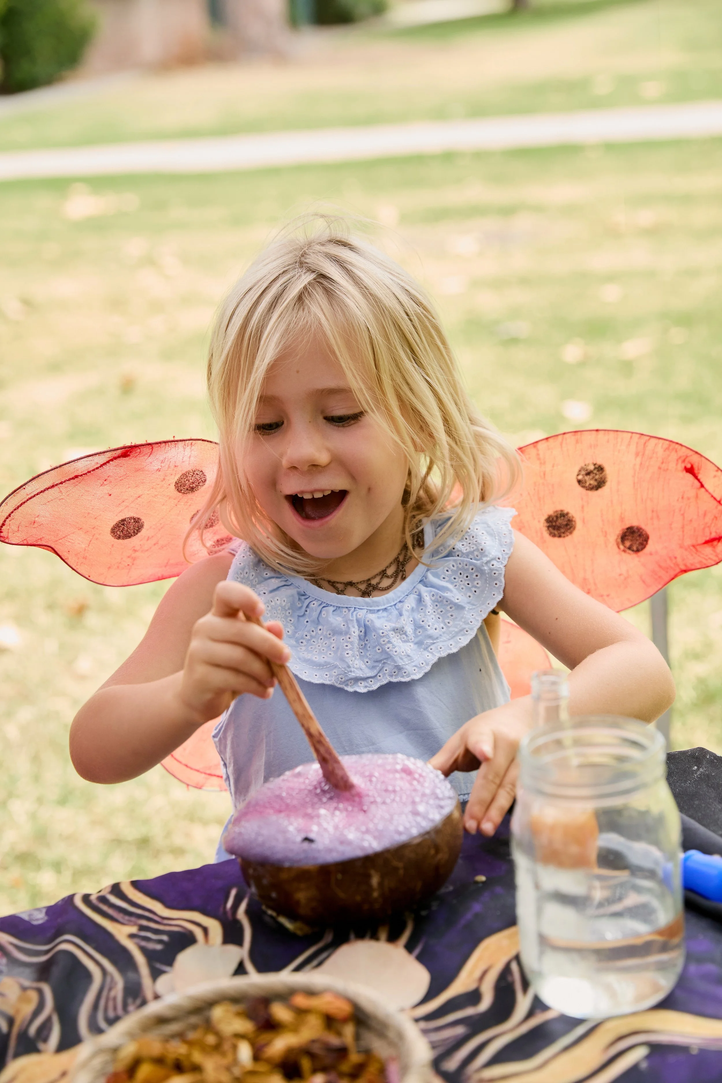 A smiling young girl wearing orange fairy wings enthusiastically mixing a potion in a coconut shell bowl.