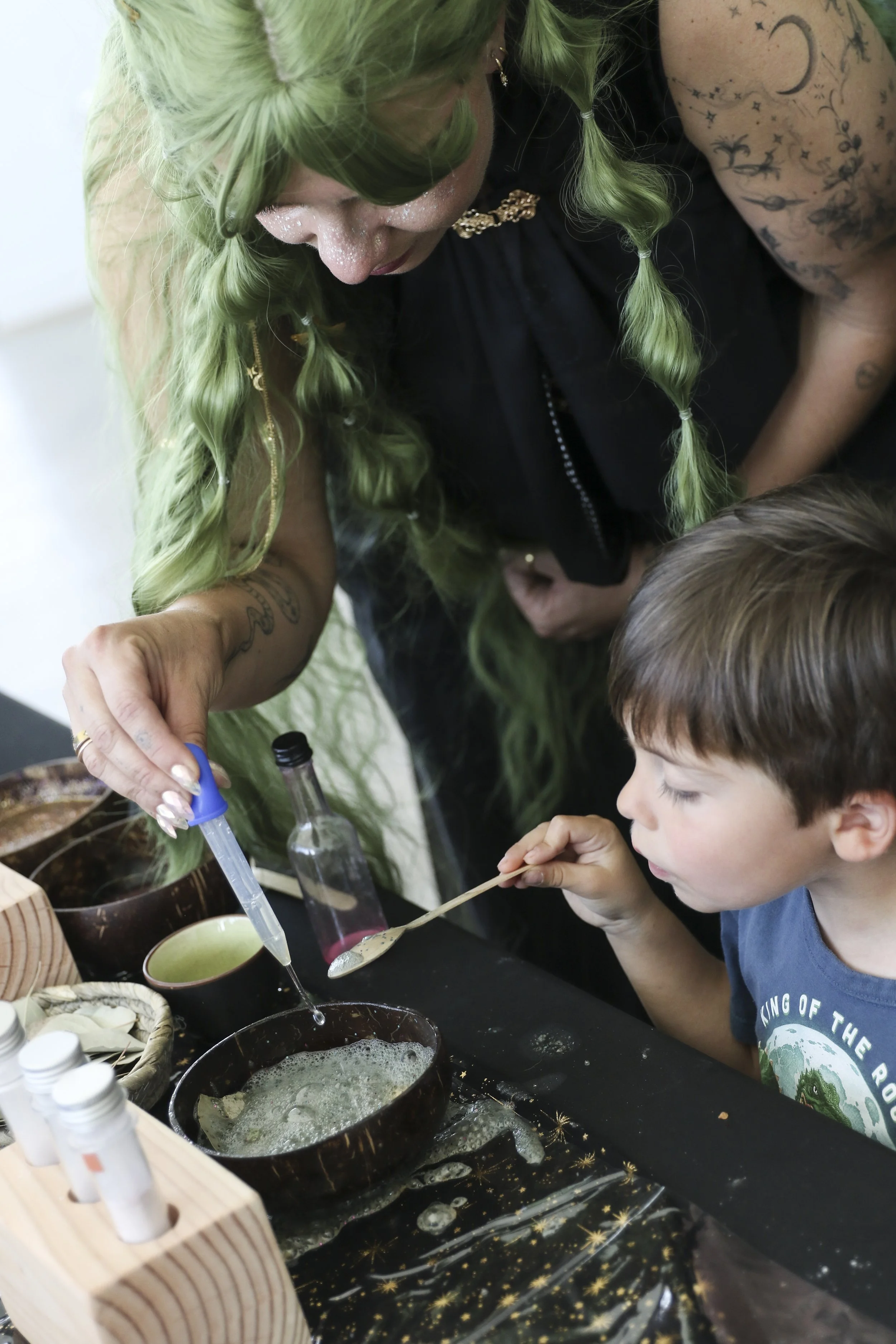 An Alchemy Guide and a young Boy engaging in a hands-on activity, using a pipette to add liquid to a foaming "potion" mixture in a coconut shell